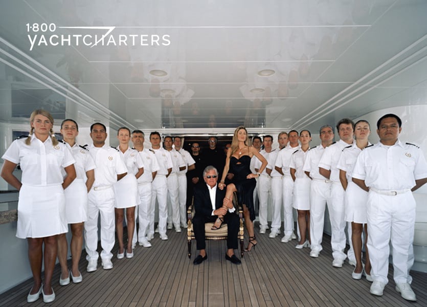 Photograph of superyacht crew in white outfits and standing at ease around owners of yacht who are wearing black. Man seated and woman in black dress standing over him. Crew in a v-formation around them.